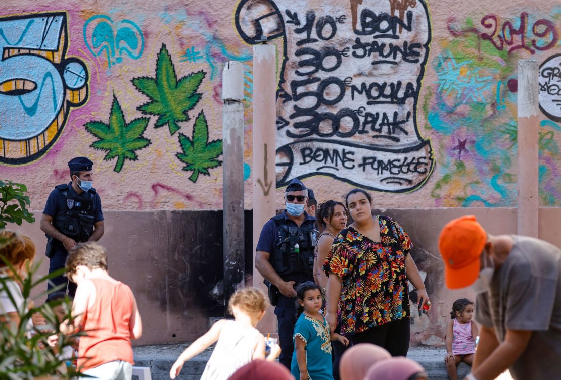 Local inhabitants and police officers stand in front of a wall in Marseille displaying prices of drugs during a visit by President Macron (unseen) on September 1, 2021. 