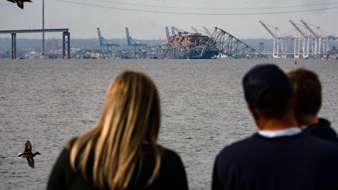 A family looks at the steel frame of the Francis Scott Key Bridge sitting on top of the container ship Dali after the bridge collapsed in Baltimore, Maryland, on March 26, 2024. The bridge collapsed early March 26 after being struck by the Singapore-flagged Dali container ship, sending multiple vehicles and people plunging into the frigid harbor below. There was no immediate confirmation of the cause of the disaster, but Baltimore's Police Commissioner Richard Worley said there was "no indication" of terrorism. (Photo by Kent Nishimura / AFP) (Photo by KENT NISHIMURA/AFP via Getty Images)