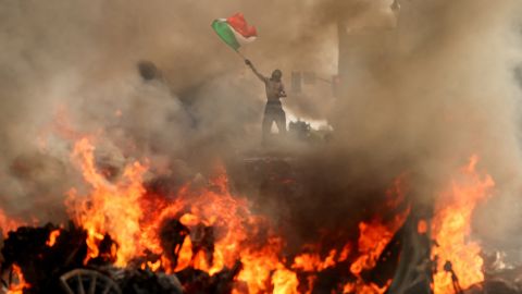 A man waves a Mexican flag as smoke and flames rise from a burning vehicle during protests in downtown Los Angeles on Sunday, June 8.