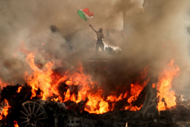A man waves a Mexican flag as smoke and flames rise from a burning vehicle during protests in downtown Los Angeles on Sunday, June 8.