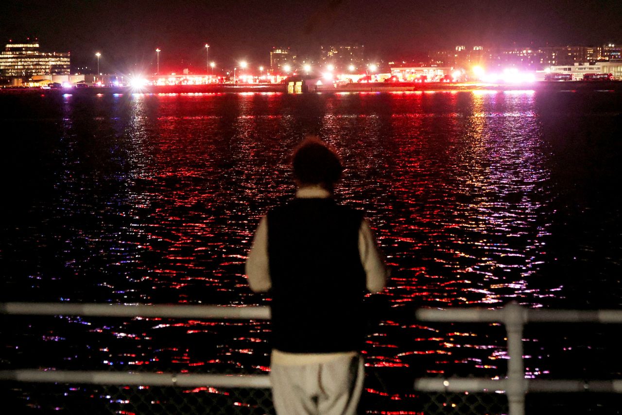 A person looks on from the bank of the Potomac River as emergency service vehicles operate near the site of the crash.