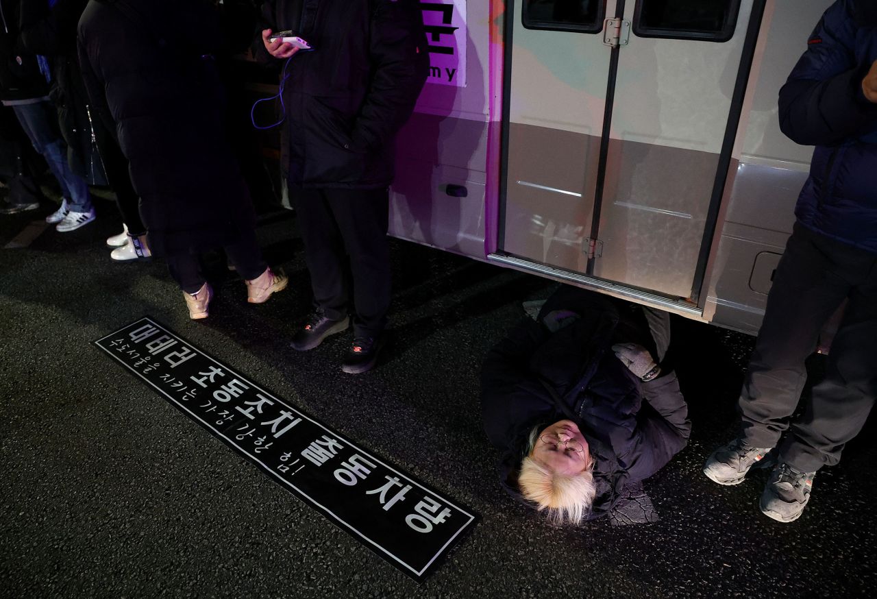 A woman lies on a road to block an army transport vehicle.