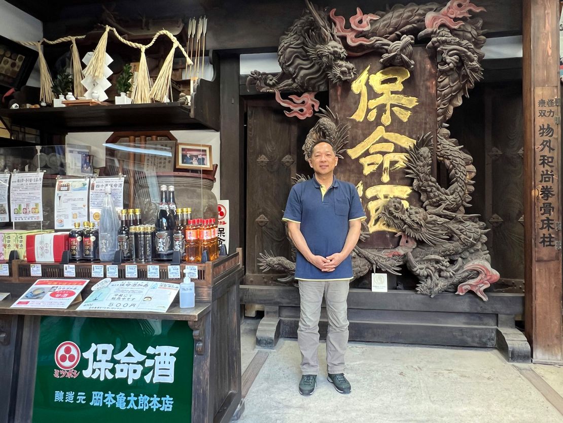 Sixth-generation brewer Ryochi Okamoto stands in front of a dragon signboard from the village's first homeishu brewer.