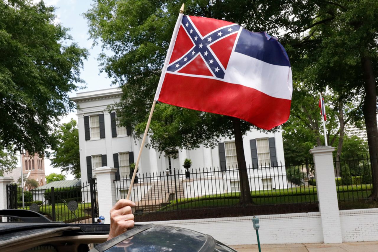 In this April 25 photograph, a small Mississippi state flag is held by a participant during a drive-by "re-open Mississippi" protest past the Governor's Mansion, in the background, in Jackson, Mississippi.