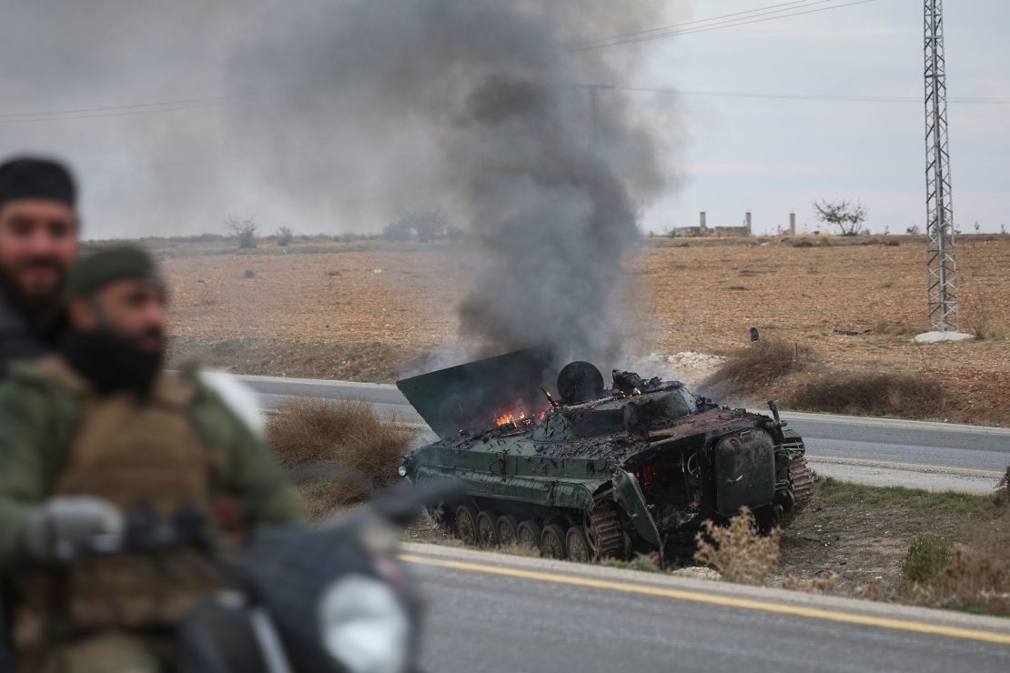 Syrian opposition fighters drive past a burning armored vehicle south of Hama, Syria, on December 7, 2024.