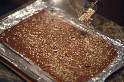 Use a scoop to sprinkle chopped pecans on top of the chocolate-covered toffee.