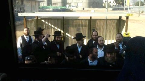 A crowd of ultra-orthodox men gather at a bus stop while Tanya Rosenblit refuses to sit in the back of the bus.