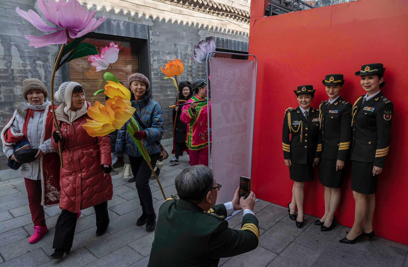 Female military officers take photos as local residents carry large flowers during a small procession for the Little Year, or Xiaonian, which marks the beginning of preparations for the Lunar New Year and Spring Festival, in Beijing, on Wednesday.