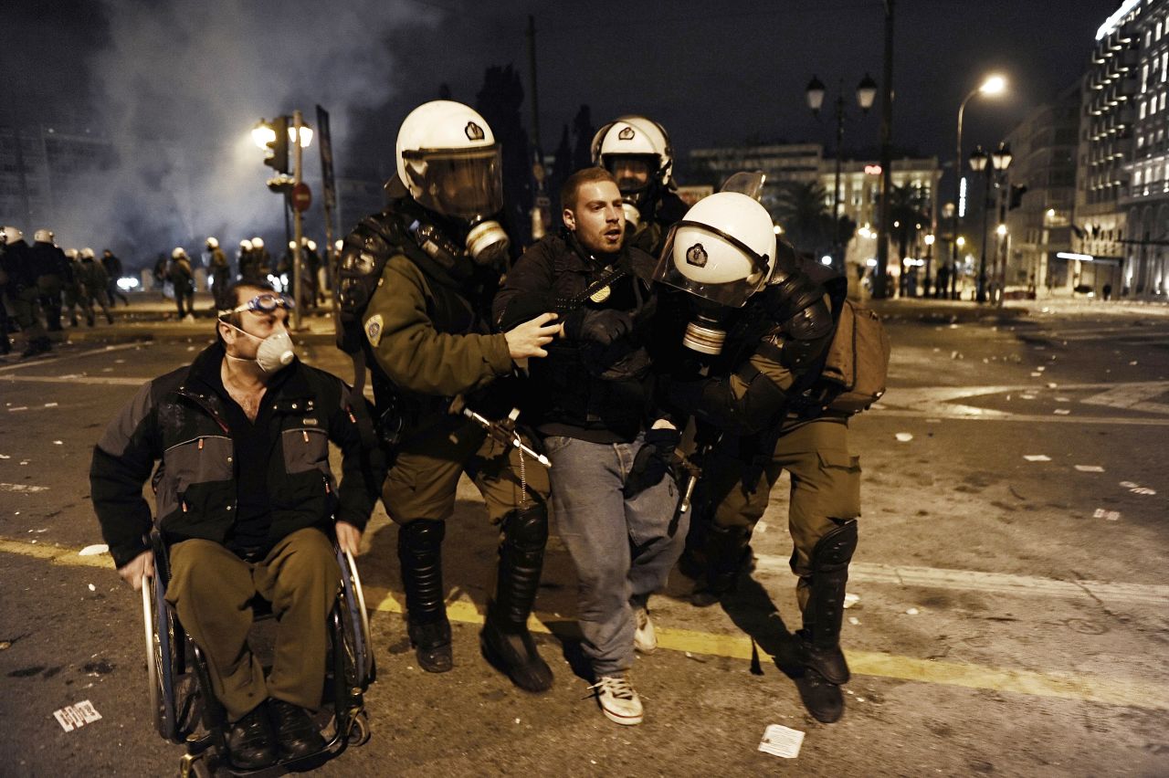 A protester is arrested and taken away by riot police while a man in a wheelchair watches. 