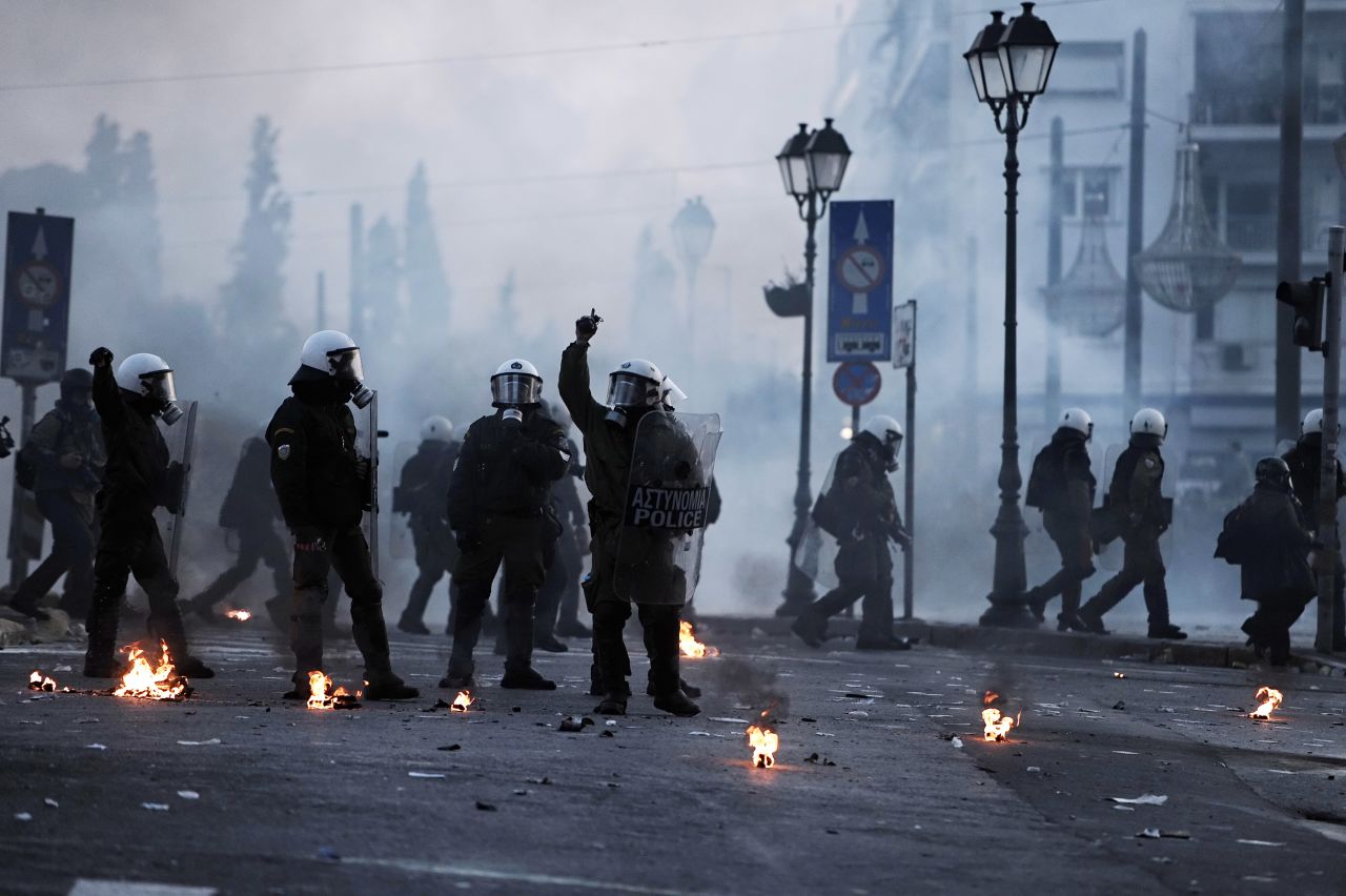 Tear gas and smoke create an eerie, fog-like atmosphere as masked riot police stand guard. 