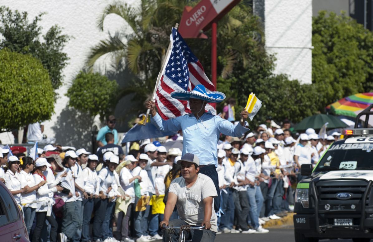 A man waves national flags of the United States and Vatican City as young people wait on the sidewalk for the arrival of the pope along Lopez Mateos Boulevard.