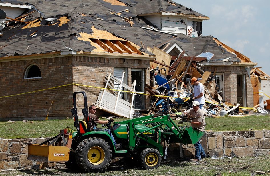 Volunteers use a small tractor to clear debris in front of a damaged house.
