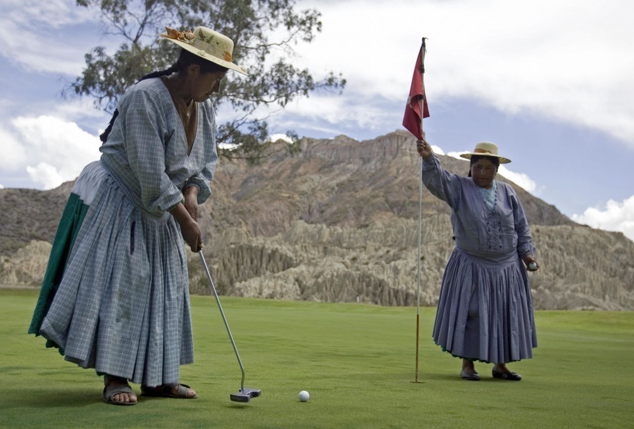 An  indigenous Aymara woman plays an approach shot as her colleague holds the flag.