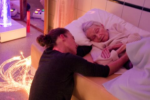 Jacqueline, a French Alzheimer's patient, needs help getting into bed. Caregiver Cossève wraps her in a soft blanket. <a href=