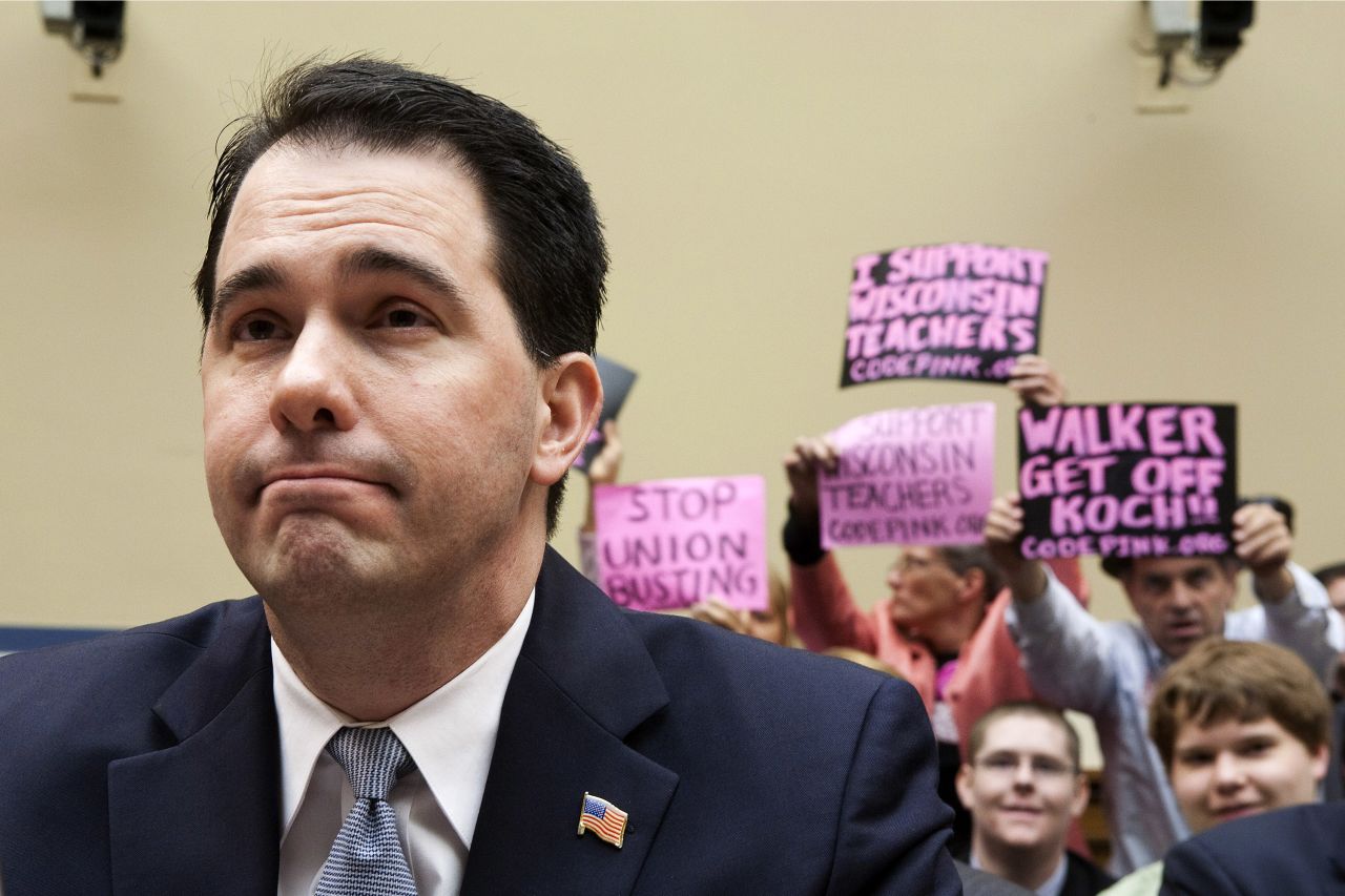 Walker testifies at a House Oversight and Government Reform Committee hearing on Capitol Hill in Washington as protesters wave signs behind him, April 14, 2011. 
