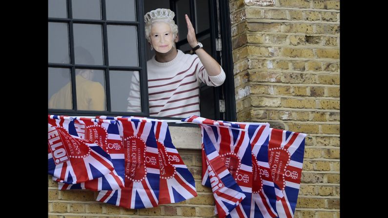 A woman wearing a mask of Queen Elizabeth II waves from a window near Tower Bridge.