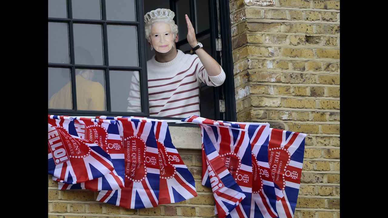 A woman wearing a mask of Queen Elizabeth II waves from a window near Tower Bridge. 
