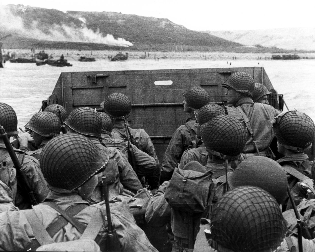 U.S. troops huddle behind the protective front of their landing craft as it nears a beachhead. Smoke in the background is naval gunfire, giving cover to troops on land.