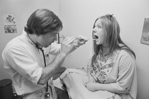 A young woman sits cross-legged on a examination table as a doctor looks inside her mouth at the Free People's Clinic in Ann Arbor, Michigan, in 1971.