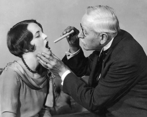 A doctor examines the throat of a young woman using a small pen light  around 1927.