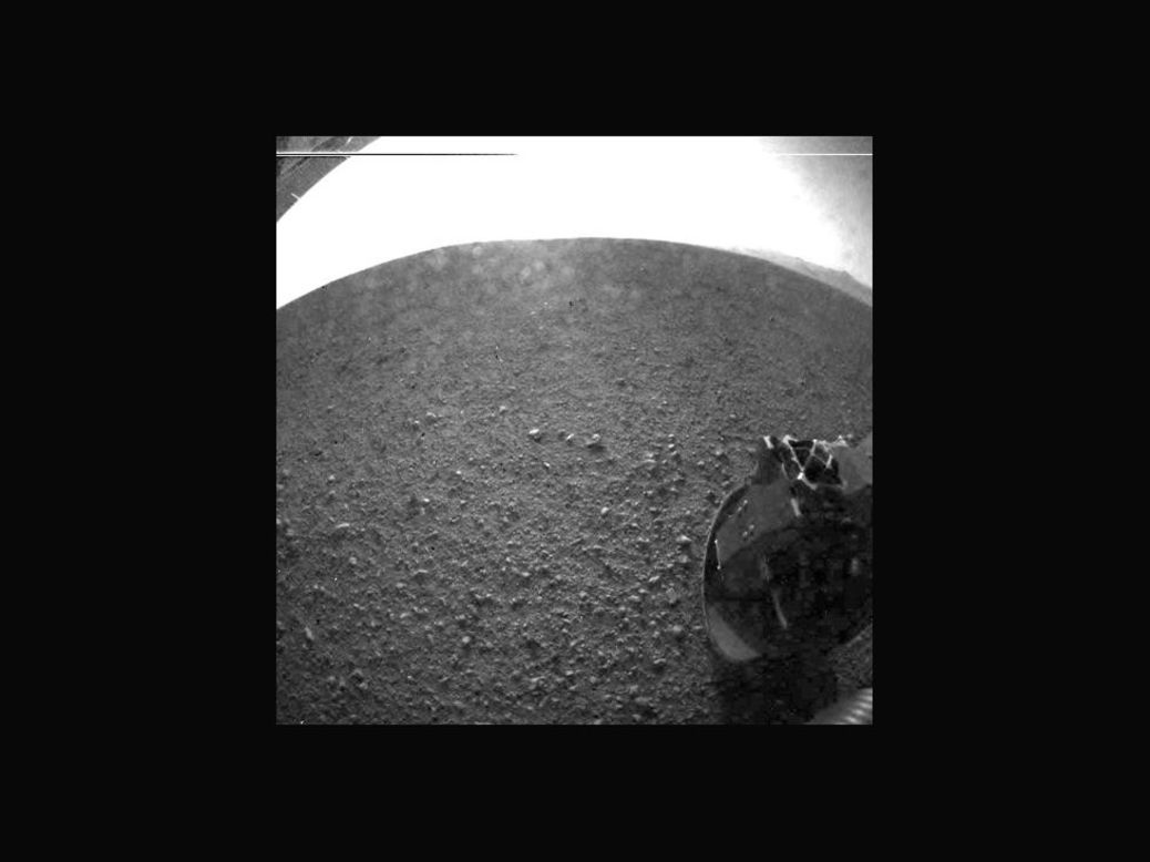 Otra de las primeras imágenes tomadas por el rover Curiosity. La cubierta de polvo claro que protegió la cámara durante el aterrizaje se abrió. Parte del resorte que liberó la cubierta de polvo se puede ver en la parte inferior derecha, cerca de la rueda del vehículo todo terreno.