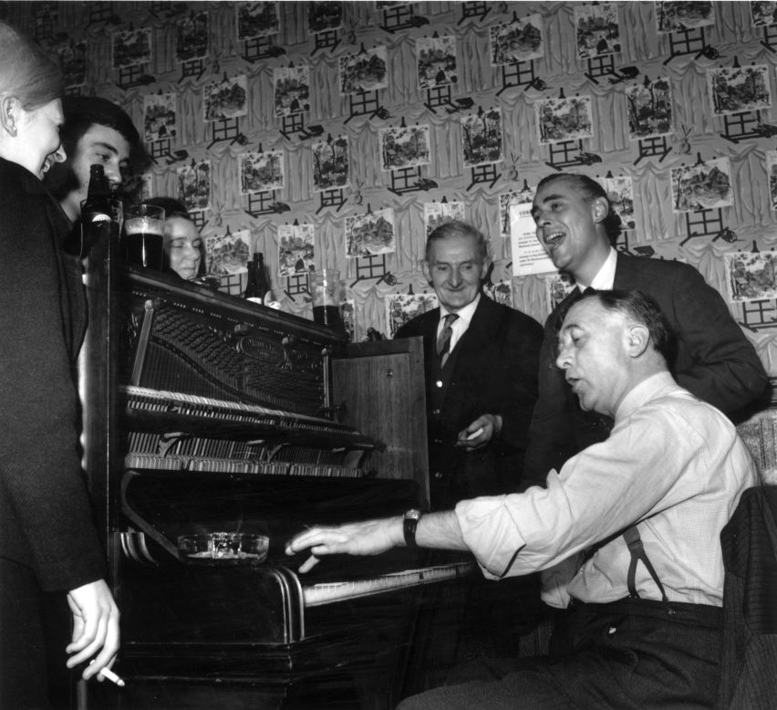 Regulars at a pub in 1964 gather round the piano for a singalong. Or to roughly translate into East London cockney: "A bunch of bacardi breezers gather down the rub-a-dub-dub for a ding dong round the old joanna." (Getty Images).