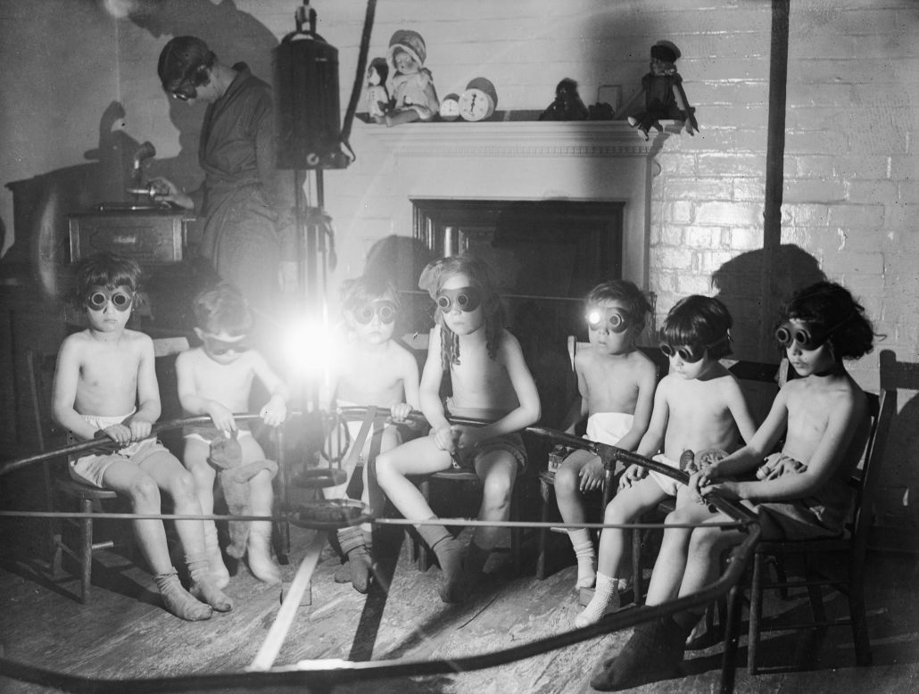 A group of children at the East End Mission on Commercial Road, Tower Hamlets in May 1931, stripped to the waist and wearing goggles, sit around a sunlamp for sun ray treatment. An adult supervisor in the background lifts the needle on a gramophone player (Getty Images).