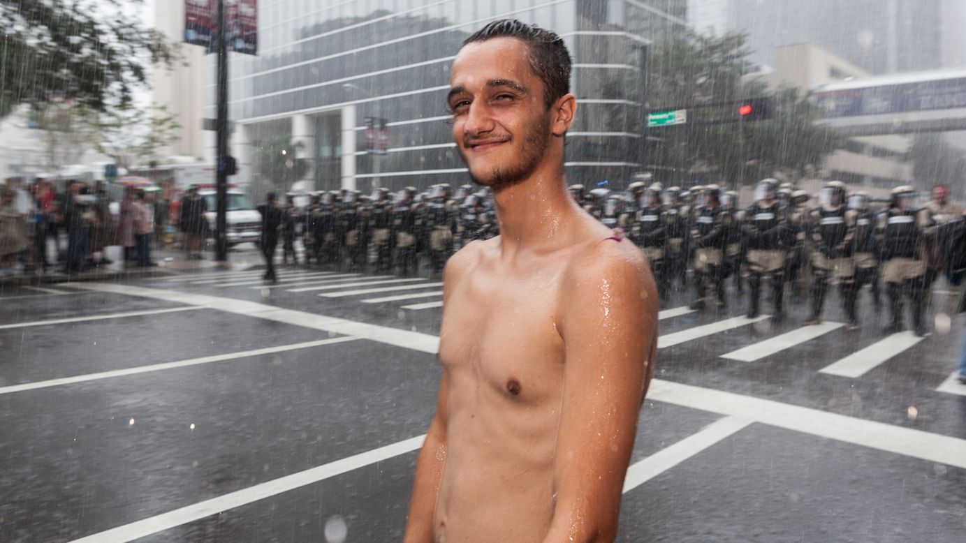 A shirtless protester stands in the rain in front of a police line.