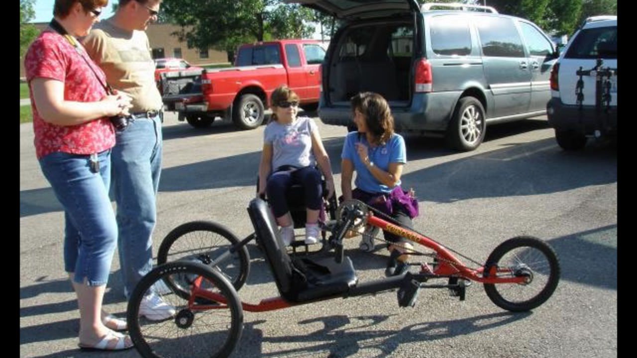 Alice Teisan has had chronic fatigue syndrome for the past 20 years. An avid cyclist who can no longer cycle, she founded His Wheels International in 2005 to help provide transportation to those in need. Here, parents Kern and Dawna Lunde watch physical therapist Nancy Biedry coach their daughter Anika, who is paralyzed from the waist down, as she learns to use her new trike prototype.