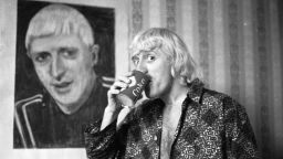 4th February 1965: Disc-jockey, Jimmy Savile poses by a portrait of himself, painted by a friend, while enjoying his regular breakfast of coke and a cigar in the Bloomsbury hotel room which he has made his home. (Photo by Chris Ware/Keystone Features/Getty Images) 