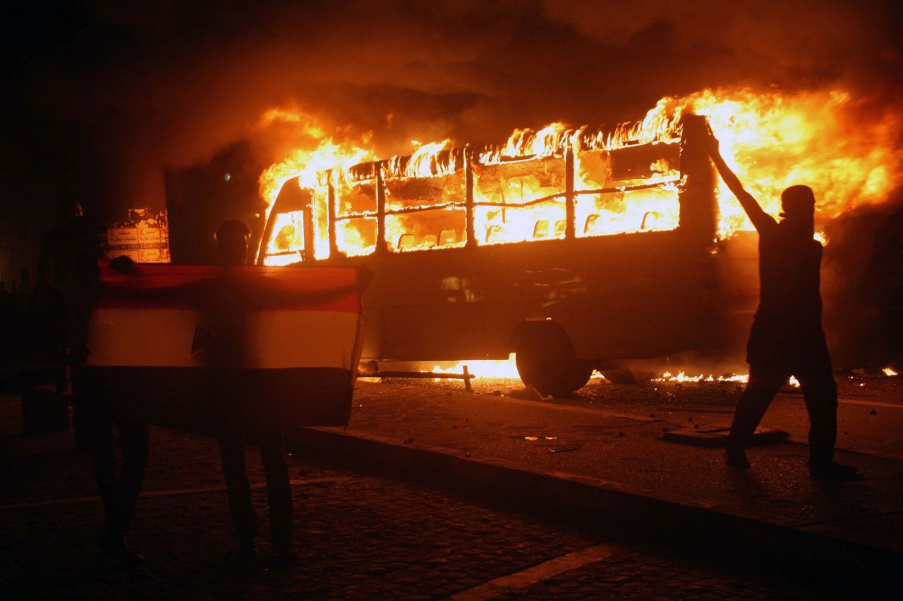 Egyptian protesters hold a national flag as they walk past a burning bus.
