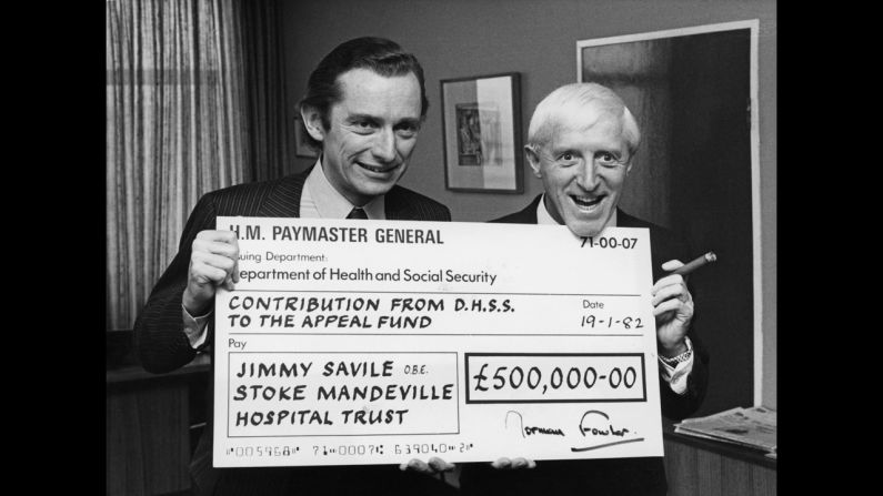 British Secretary of State for Social Services Norman Fowler presents Savile with a check for half a million pounds in 1982 as the government's contribution to an appeal for a new spinal injuries unit at Stoke Mandeville Hospital.