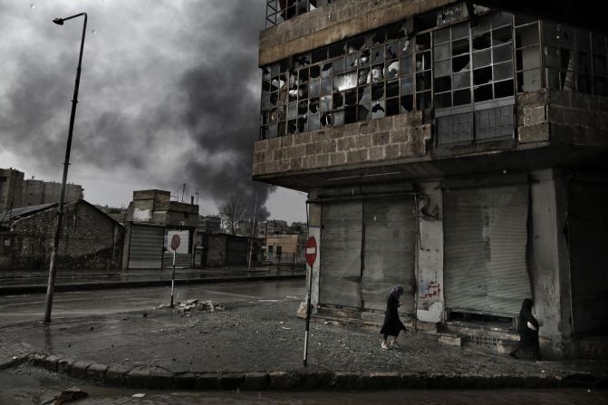 Smoke rises from a fuel station following a mortar attack as Syrian women walk on a rainy day in the Arqub neighborhood of Aleppo on October 25.