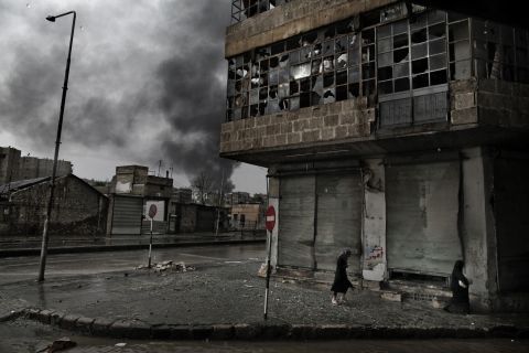 Smoke rises from a fuel station following a mortar attack as Syrian women walk on a rainy day in the Arqub neighborhood of Aleppo on October 25, 2012.