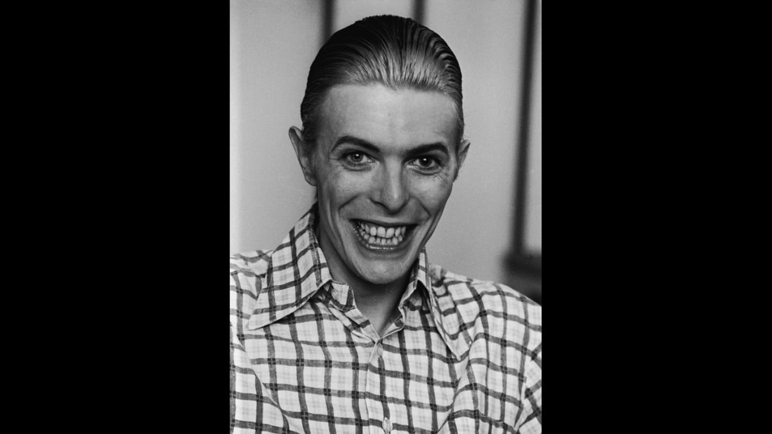 Bowie grins broadly, wearing a plaid shirt with his hair slicked back, circa 1980. 