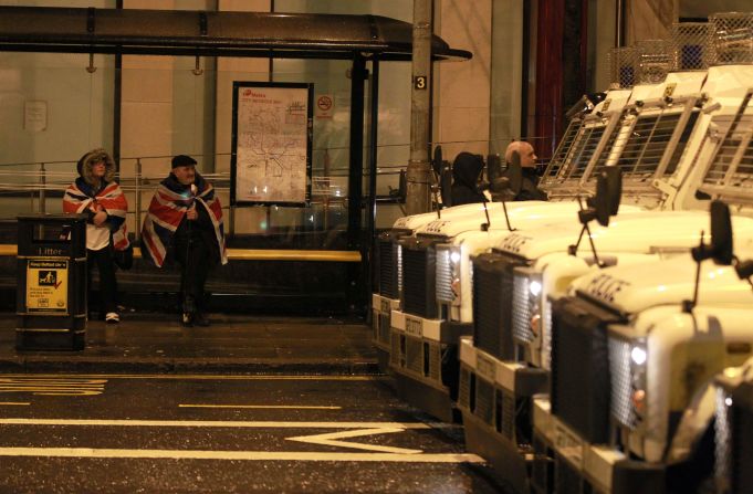 A couple sit at a bus stop wrapped in British union flags as police vehicles protect the back of the City Hall on Monday.