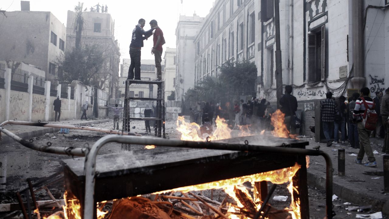 Two protesters stand on top of a piece of furniture while riot police watch from a nearby rooftop in Cairo.