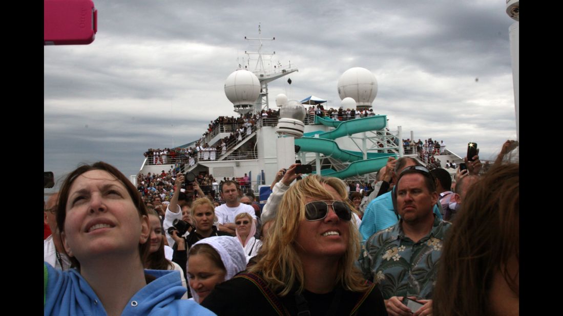 Passengers fill the deck of the ship and look up toward the sky. 