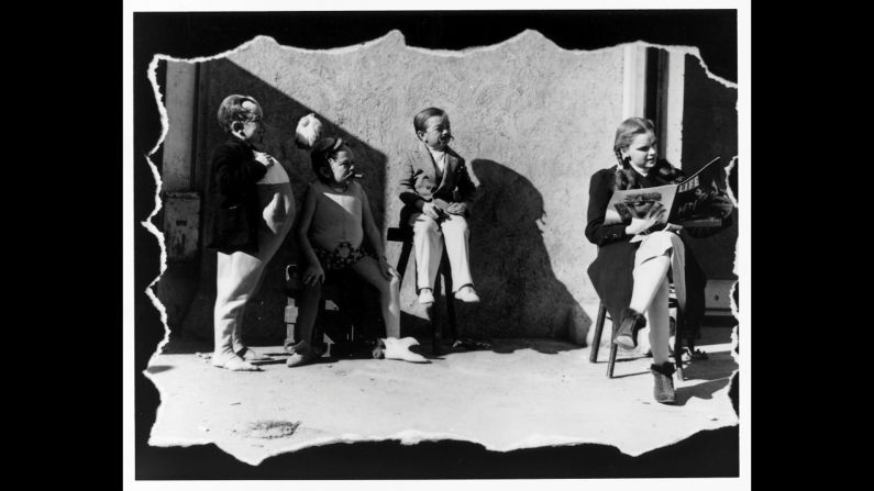 Judy Garland reads a copy of Life magazine on set as three of the actors playing Munchkins stand by.