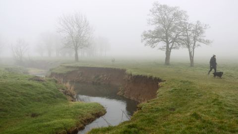 Fog shrouds a dog and its owner as they walk beside Bourne Brook, in Toft Cambridgeshire, United Kingdom, on Friday, March 8.