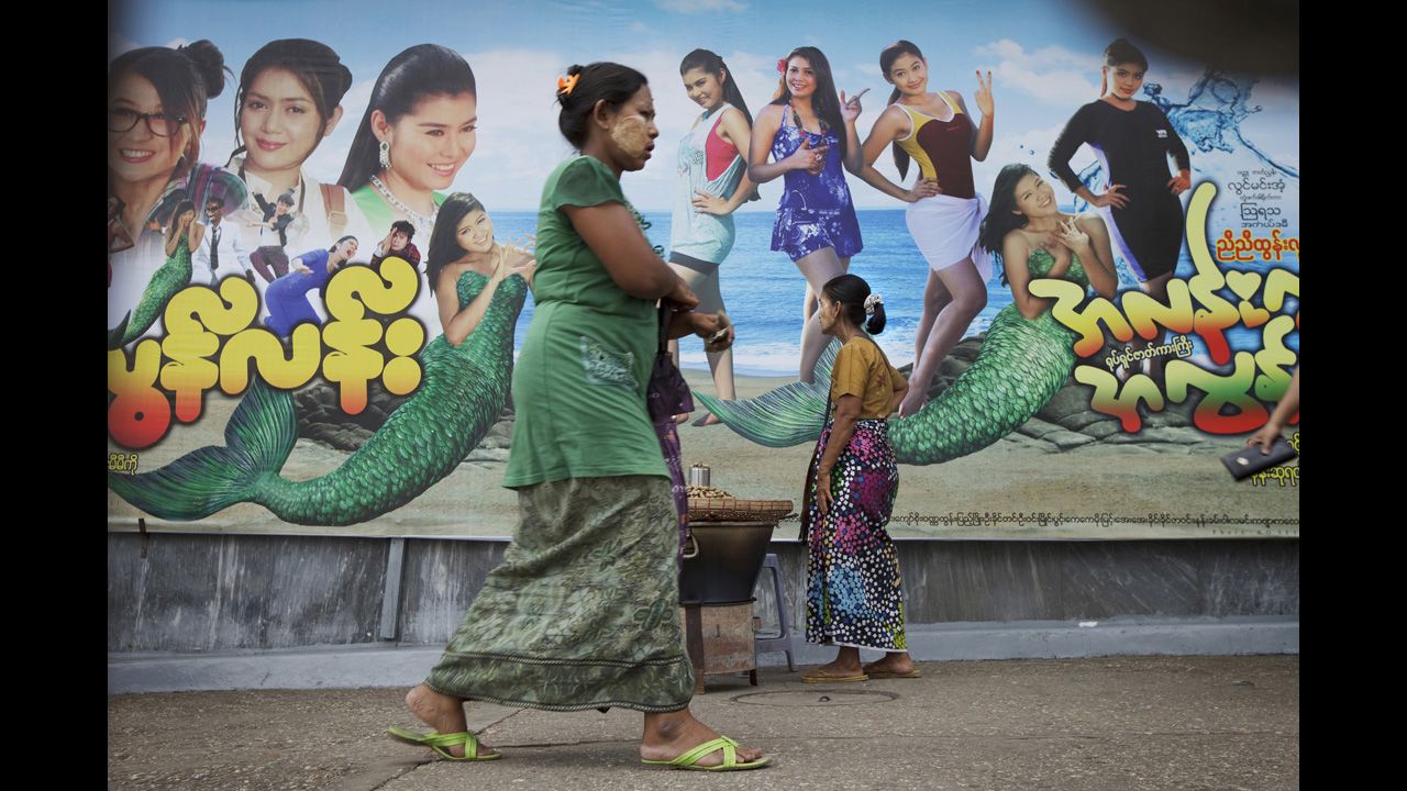 Women wear traditional longyi around their waists in front of a modern movie billboard in Yangon.