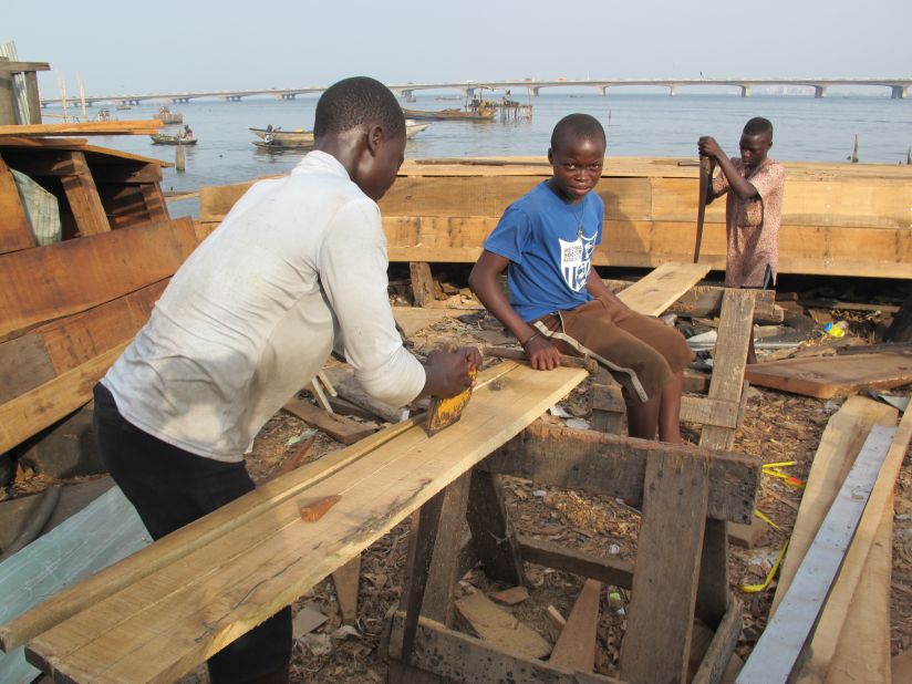 Young men in Makoko are typically put to work building canoes, in business ventures supervised by elders. Here, one teen sits on a plank of timber as two others work to saw it into pieces.