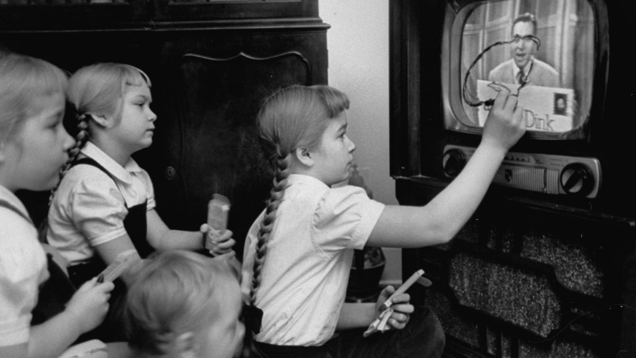 A girls uses her "Winky Dink" drawing kit to draw on a TV screen as they watch the 1950s kids program.