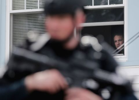 A man watches from the window of a home as a SWAT team member keeps watch on Friday, in Watertown, Massachusetts.