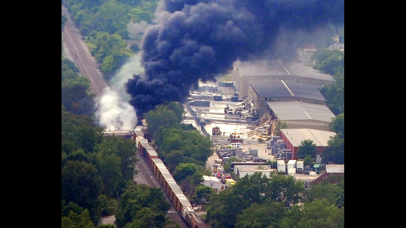 Several of the train cars are off the track as white and black smoke rises in separate columns from the wreckage.