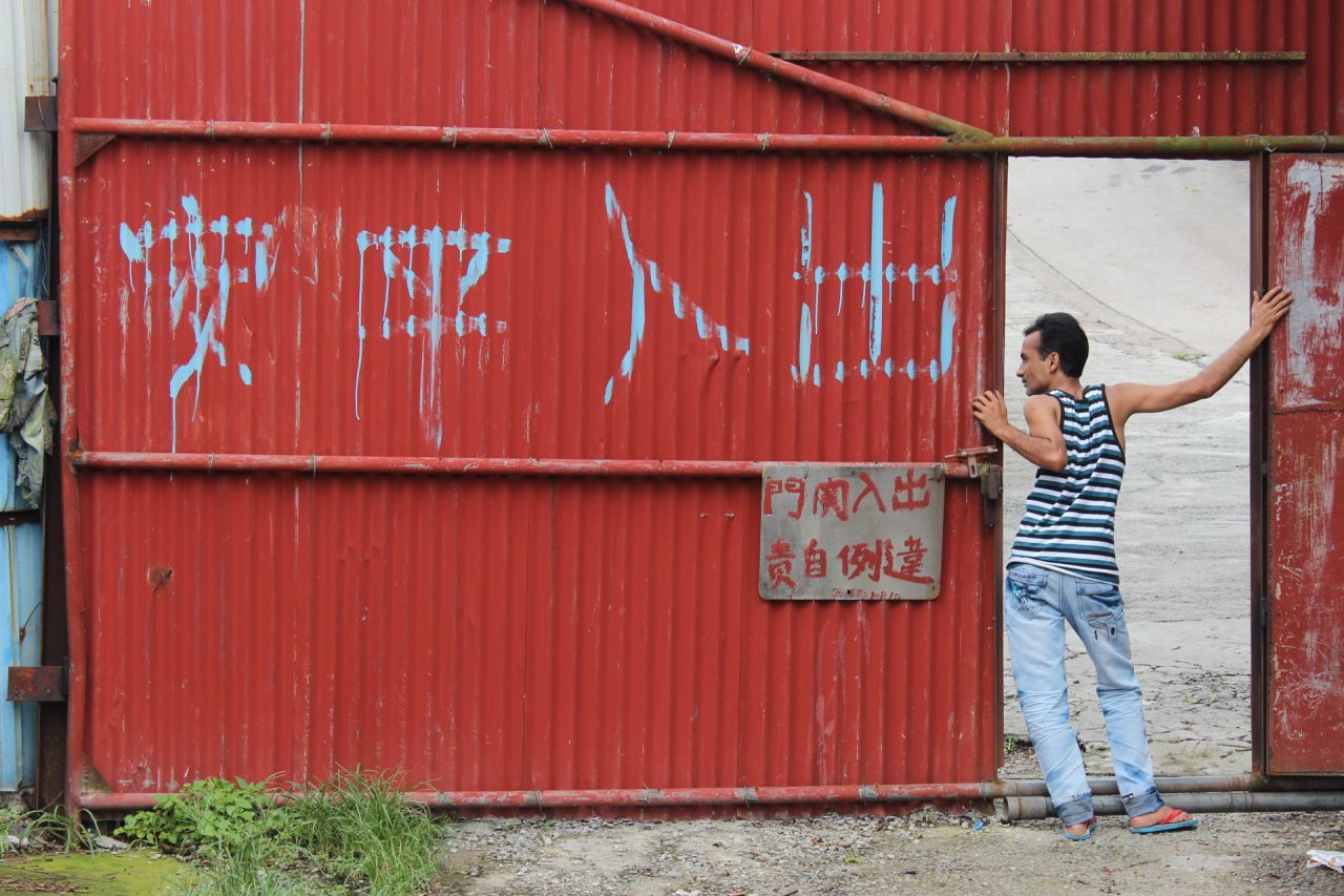 Delwar, a refugee from Bangladesh, leans out of the main entrance of a housing compound in the slum village of Ping Che on June 25, 2013. The large Chinese characters read, "Peace and safety to those who enter and leave."