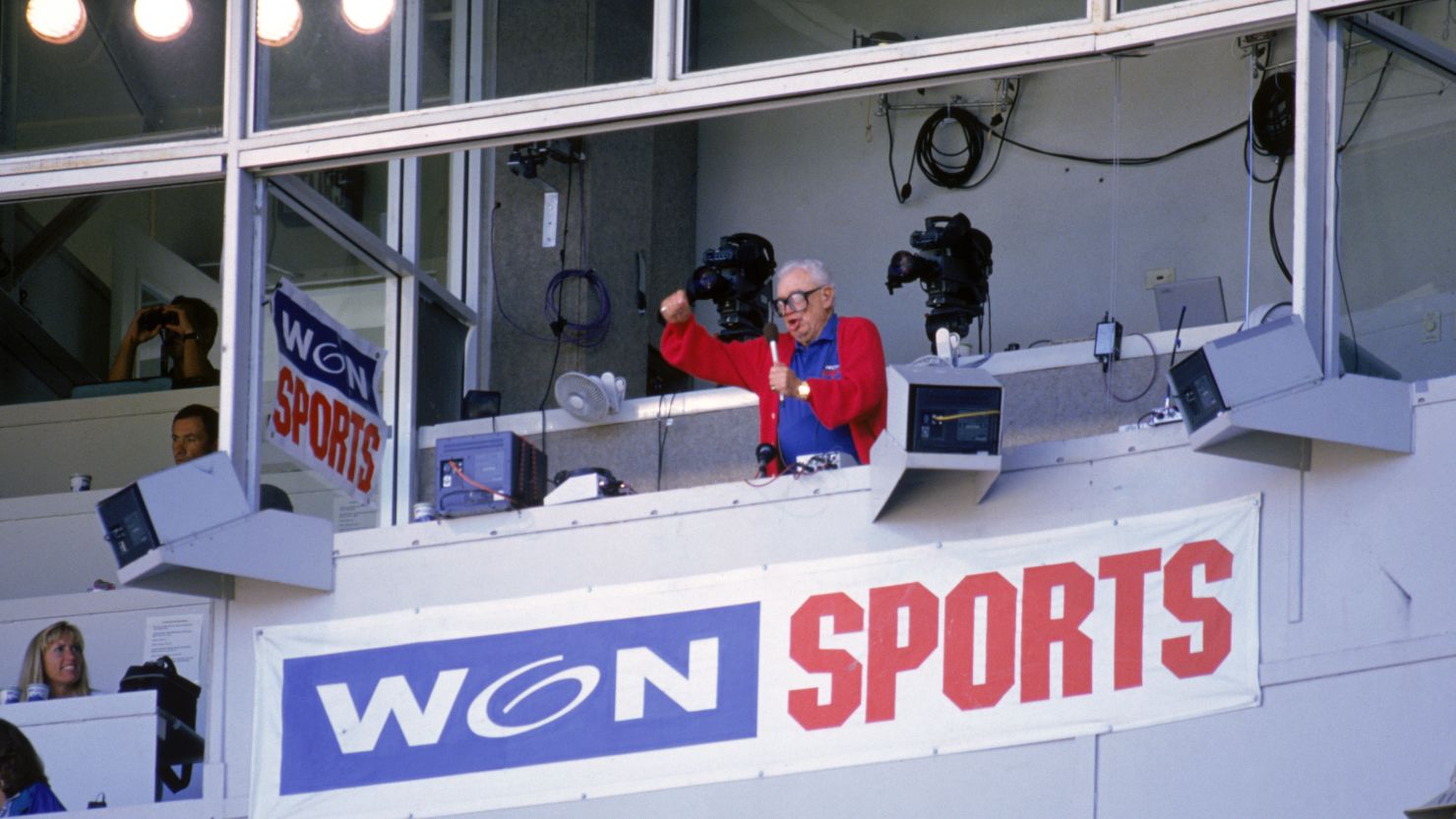 Chicago Cubs baseball announcer Harry Caray conducts fans singing "Take Me Out to the Ball Game" from his television booth in 1996