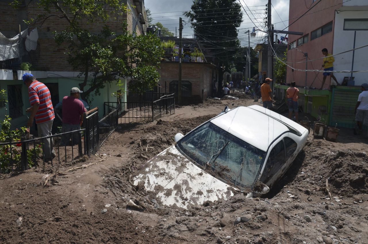 A car lies submerged in mud as residents attempt to clean up.