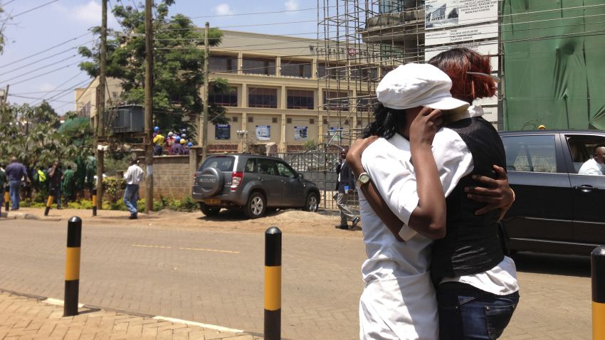 Two women hug outside an upscale shopping mall, seen background left , in Nairobi, Kenya Saturday Sept. 21 2013, after shooting erupted when armed men attempted to rob a store. Bursts of gunfire were heard from outside the mall Saturday, where cars were left abandoned. Witnesses say a half dozen grenades also went off along with lobbies of gunfire that started at midday. Police say they are engaging the attackers. (AP Photo/ Jason Straziuso)