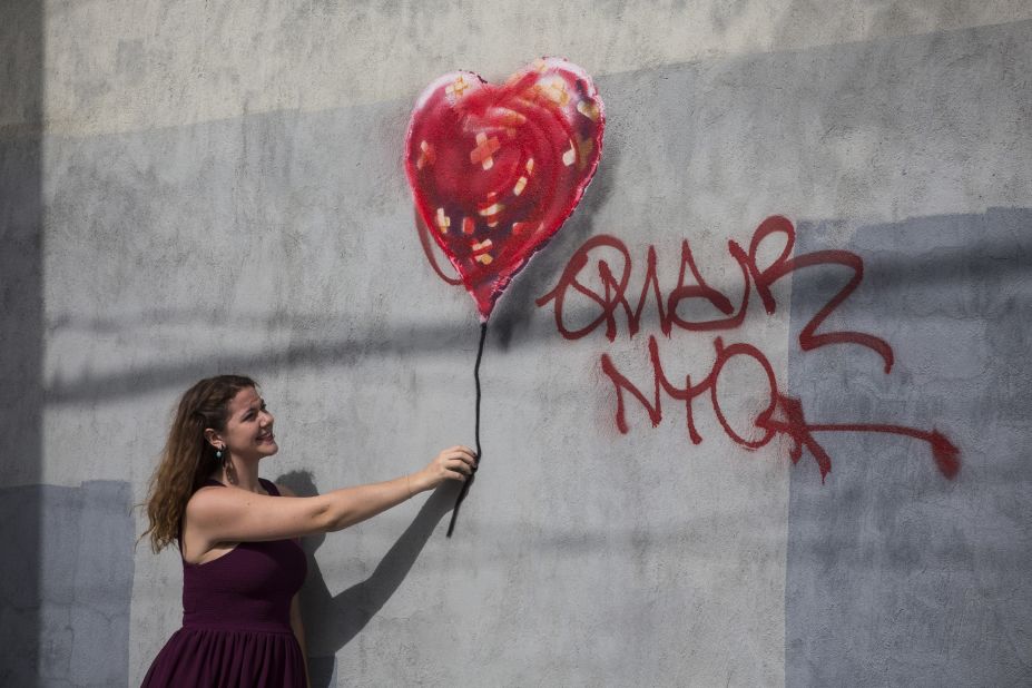 A woman poses with Banksy's painting of a heart-shaped balloon covered in bandages. The piece, in the Red Hook neighborhood of Brooklyn, was defaced with red spray paint shortly after it was completed.